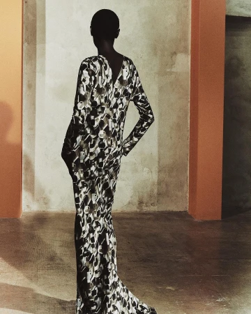 Woman poses indoors in a long-sleeved, patterned gown. She is facing away from the camera with an indoor concrete background.
