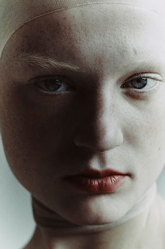 Extreme close-up portrait of a woman with pale skin, freckles, light eyebrows, and reddish lips wearing a cap.