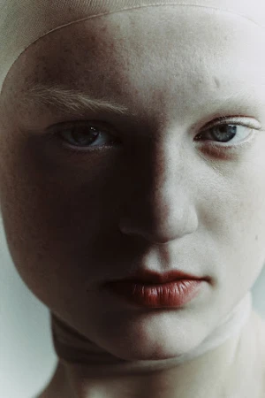Extreme close-up portrait of a woman with pale skin, freckles, light eyebrows, and reddish lips wearing a cap.