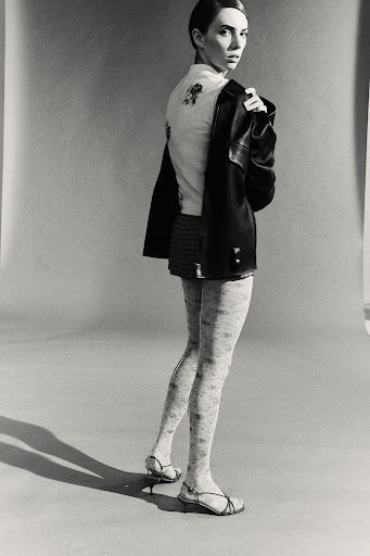 A woman poses indoors, wearing a jacket and skirt. She has light skin, dark hair, and sandals, looking toward the camera.