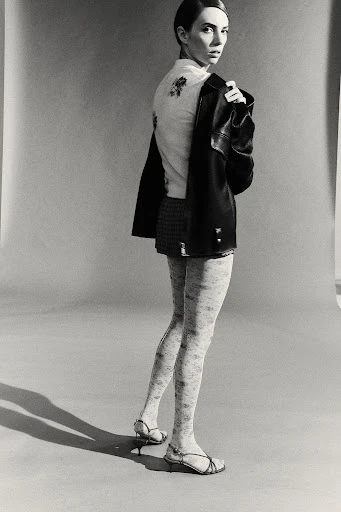 A woman poses indoors, wearing a jacket and skirt. She has light skin, dark hair, and sandals, looking toward the camera.