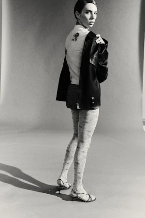 A woman poses indoors, wearing a jacket and skirt. She has light skin, dark hair, and sandals, looking toward the camera.