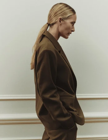 Profile portrait of a blonde woman wearing an olive green blazer and matching trousers against a pale wall.