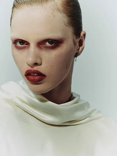 Close-up portrait of a woman with red hair and heavy red eye makeup. She is wearing a white top with a high neck.