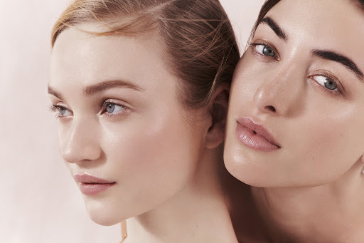 Close-up studio shot of two fair-skinned adult women with glossy skin, posing intimately in front of a blurred background.