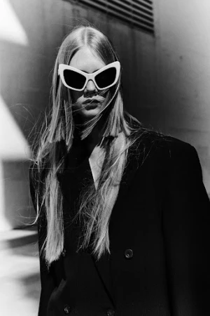 Model with long blonde hair wearing oversized white sunglasses and a black coat against a textured wall.