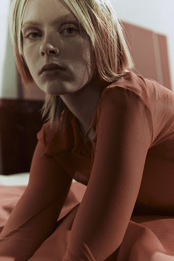 Woman with blonde hair and red top sitting on a red surface indoors looking at the camera in a medium shot, with red walls in the background.