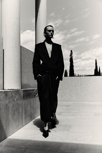 Model in a black blazer and long skirt walking past tall concrete columns under a bright sky.