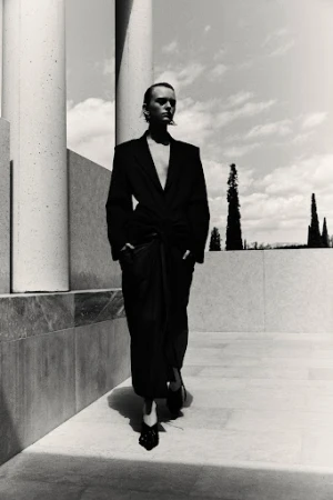 Model in a black blazer and long skirt walking past tall concrete columns under a bright sky.
