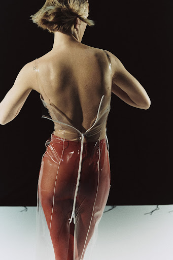 Back view of woman in a transparent top with a zipper and brick-red pants posing in a studio with a dark background.