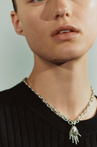 Close-up of a fair-skinned young adult with a silver chain necklace and hand-shaped pendant against a plain light background.