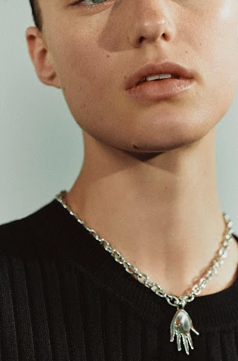 Close-up of a fair-skinned young adult with a silver chain necklace and hand-shaped pendant against a plain light background.