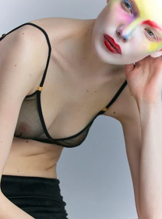 Model wearing a black sheer bra with colorful face paint and bold red lips against a neutral background.