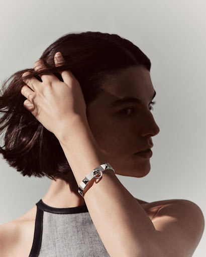Woman poses in a studio with hand in her hair and a white and silver bracelet, in a beauty shot, against a light background.