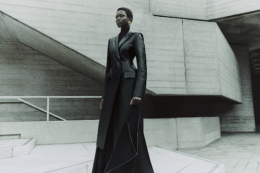 Adult woman in a dark coat posing on stone steps in front of a textured concrete wall, creating an editorial fashion portrait.