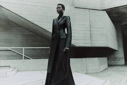 Adult woman in a dark coat posing on stone steps in front of a textured concrete wall, creating an editorial fashion portrait.