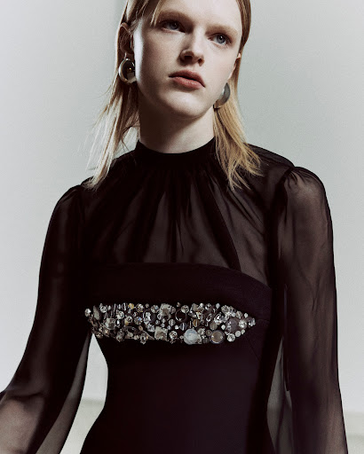 Close-up of a blonde adult female wearing a black gown with jewel embellishments, captured in a photo studio setting with neutral lighting.