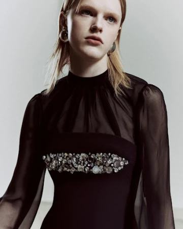Close-up of a blonde adult female wearing a black gown with jewel embellishments, captured in a photo studio setting with neutral lighting.