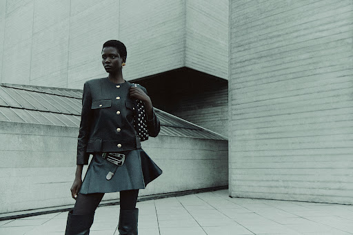 Fashionable woman poses in front of modern concrete architecture wearing a jacket, skirt, tights, and boots, with a studded purse.