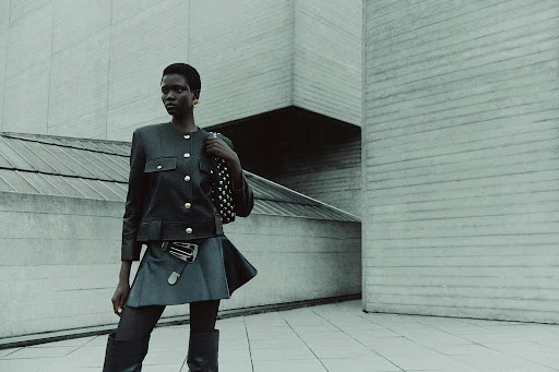Fashionable woman poses in front of modern concrete architecture wearing a jacket, skirt, tights, and boots, with a studded purse.