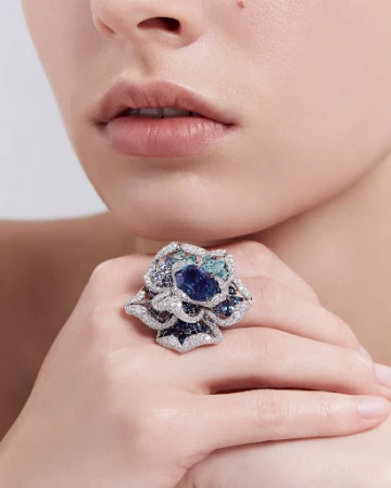 Close-up of a woman's hand wearing a large flower-shaped statement ring with blue and green gems and diamonds against a light background.