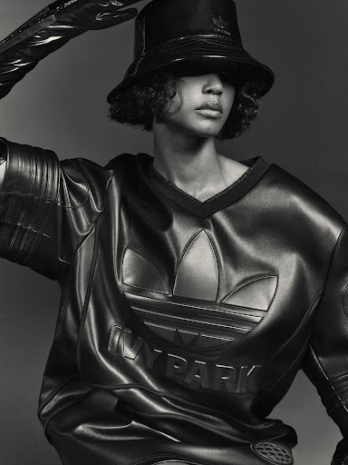 Woman in black bucket hat and leather outfit posing in studio with hand raised. Adidas and Ivy Park logos are visible on clothing.