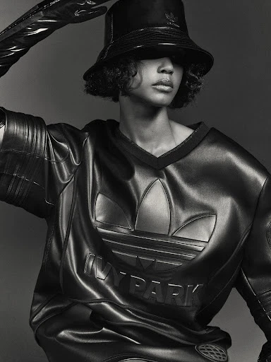 Woman in black bucket hat and leather outfit posing in studio with hand raised. Adidas and Ivy Park logos are visible on clothing.