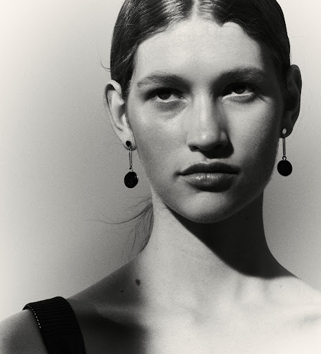Monochromatic portrait of a young adult woman with dangling black disc earrings, minimalist style, against a neutral grey backdrop.