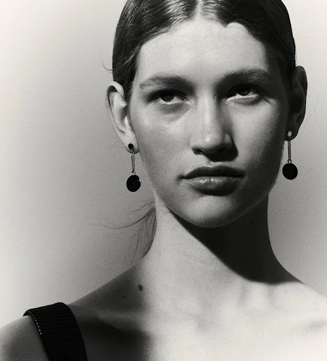 Monochromatic portrait of a young adult woman with dangling black disc earrings, minimalist style, against a neutral grey backdrop.