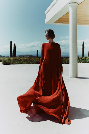 Elegant woman in flowing orange gown poses against a backdrop of modern architecture and soft, rolling hills under a clear blue sky.