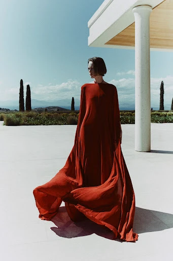 Elegant woman in flowing orange gown poses against a backdrop of modern architecture and soft, rolling hills under a clear blue sky.