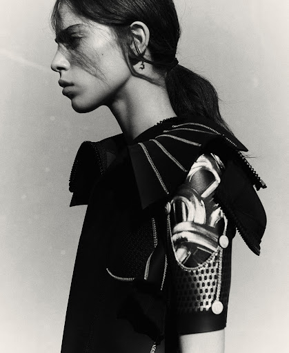 Monochrome studio portrait of a young adult woman with dark hair in a ponytail, wearing avant-garde black clothing.