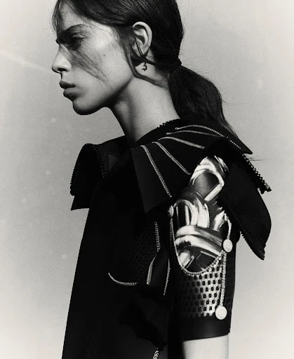 Monochrome studio portrait of a young adult woman with dark hair in a ponytail, wearing avant-garde black clothing.