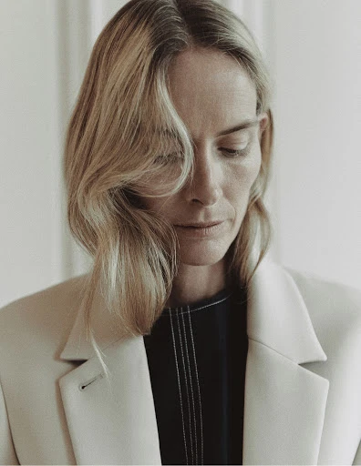Portrait of a blonde woman with fair skin wearing a cream blazer and dark top against a neutral backdrop, looking down with a gentle expression.