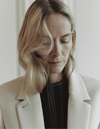 Portrait of a blonde woman with fair skin wearing a cream blazer and dark top against a neutral backdrop, looking down with a gentle expression.