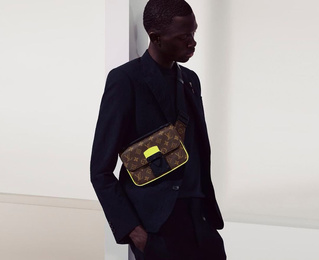 Stylish dark-skinned man in a black suit jacket, wearing a patterned Louis Vuitton messenger bag with yellow trim, against a neutral backdrop.