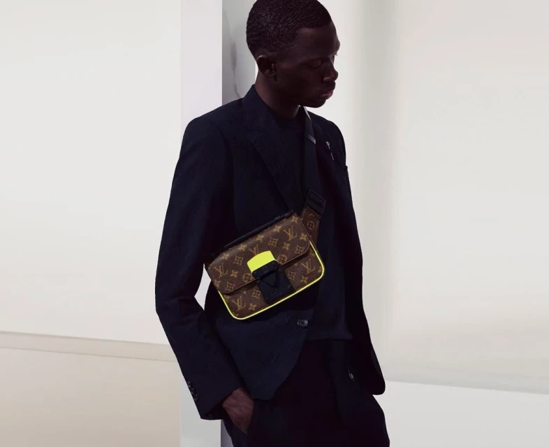 Stylish dark-skinned man in a black suit jacket, wearing a patterned Louis Vuitton messenger bag with yellow trim, against a neutral backdrop.