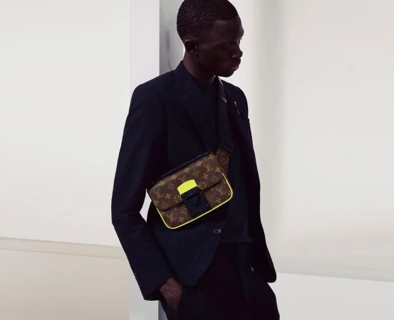 Stylish dark-skinned man in a black suit jacket, wearing a patterned Louis Vuitton messenger bag with yellow trim, against a neutral backdrop.