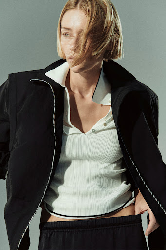 A model poses in a black jacket and off-white shirt against a grey backdrop in a fashion editorial shot with natural lighting.