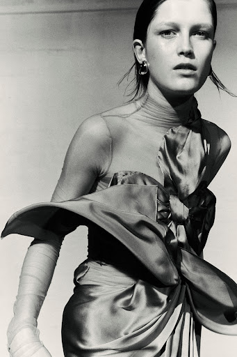 Monochromatic portrait of a woman wearing a satin dress with a large bow, complemented by sheer arm coverings and hoop earrings.