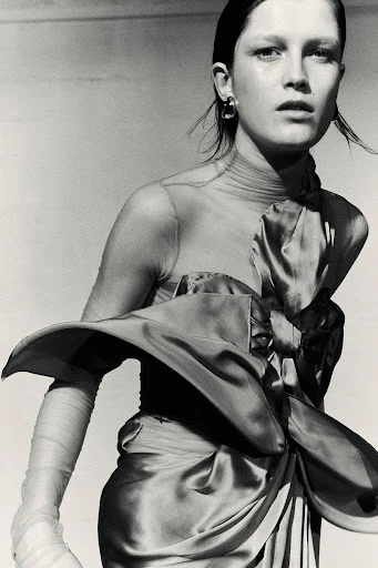 Monochromatic portrait of a woman wearing a satin dress with a large bow, complemented by sheer arm coverings and hoop earrings.