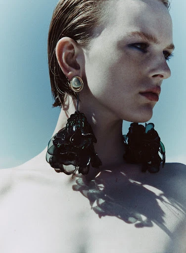 Close-up of a young adult with wet hair, wearing large dangling earrings and a jeweled stud, with a clear blue sky background.