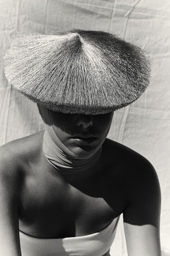 Model wearing a wide-brimmed conical straw hat casting a shadow across her face against a textured fabric background