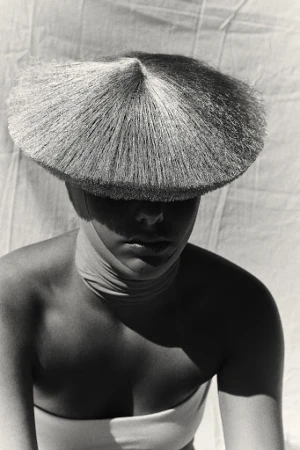 Model wearing a wide-brimmed conical straw hat casting a shadow across her face against a textured fabric background