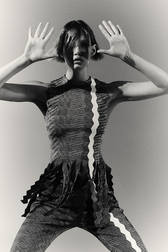 Monochromatic fashion shot of an adult female model in patterned top and pants holding her hands up, against a pale gray background.