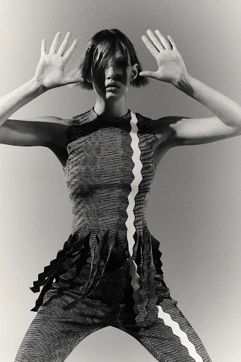 Monochromatic fashion shot of an adult female model in patterned top and pants holding her hands up, against a pale gray background.