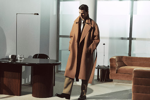 Man in a long tan coat standing in a light-filled interior. A desk and sofa provide a modern and sleek office environment.