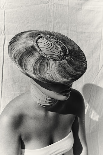 Monochrome portrait of a person wearing an elaborate sculptural hairstyle resembling a hat, styled by Melanie + Ramon