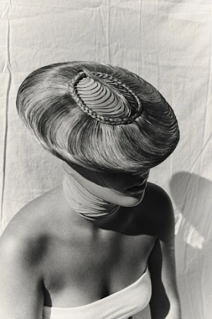 Monochrome portrait of a person wearing an elaborate sculptural hairstyle resembling a hat, styled by Melanie + Ramon