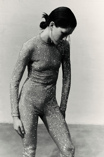 Monochrome fashion shot of a woman in a sparkling jumpsuit, long earrings, and a low ponytail against a neutral backdrop.